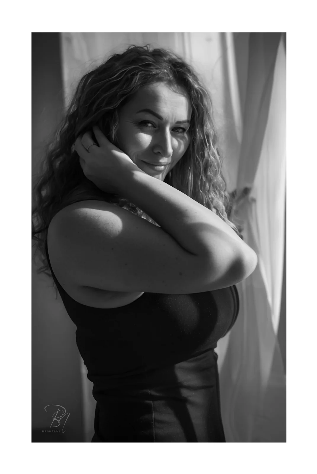 At the window — Photo: Norbert Banhalmi A woman in a black top stands by a bright window, one hand at her neck, looking away.