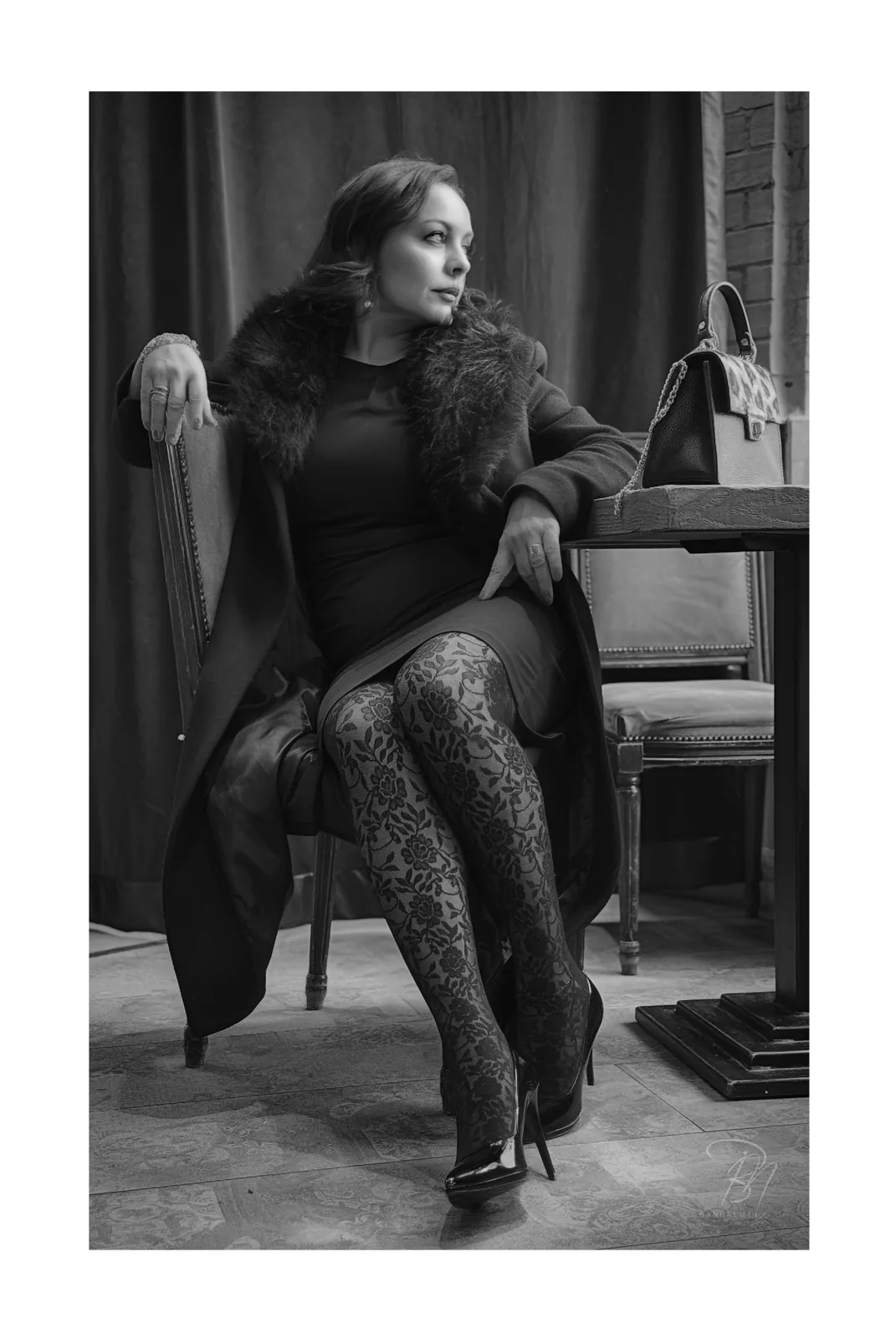 At the table — Photo: Norbert Banhalmi A woman in patterned stockings sits sideways on a chair beside a table, one hand resting behind her.