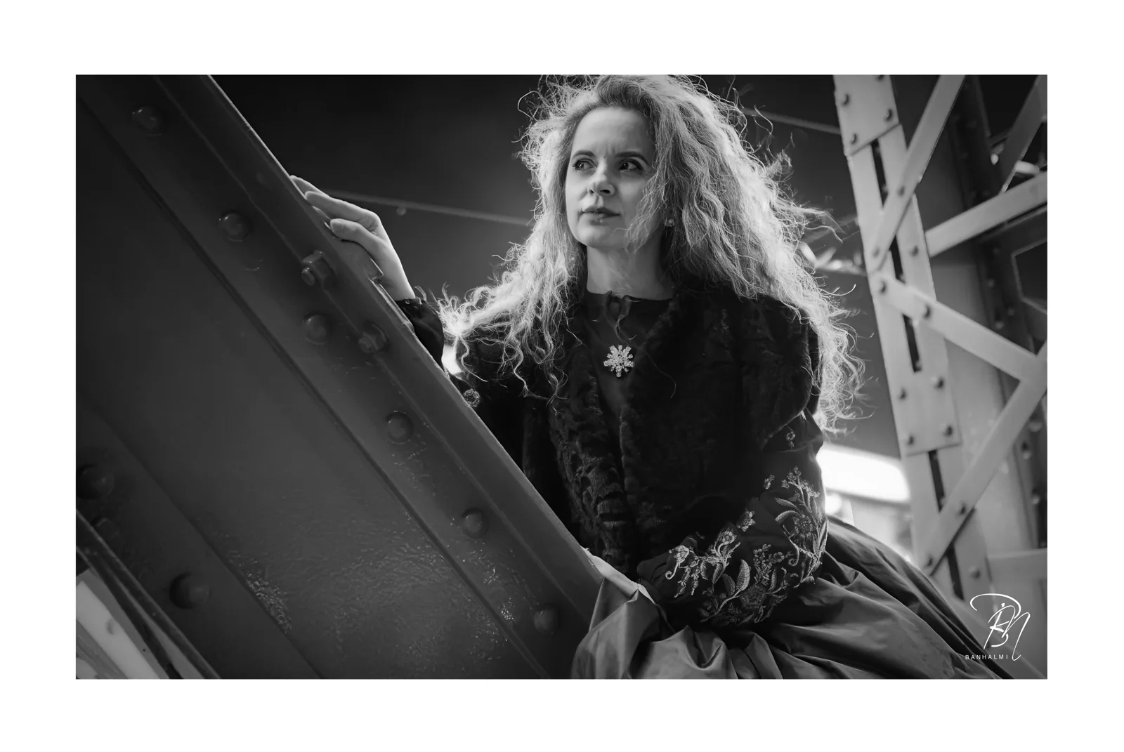 Under the iron bridge — Photo: Norbert Banhalmi A woman in a period dress leans against the riveted steel of a bridge.