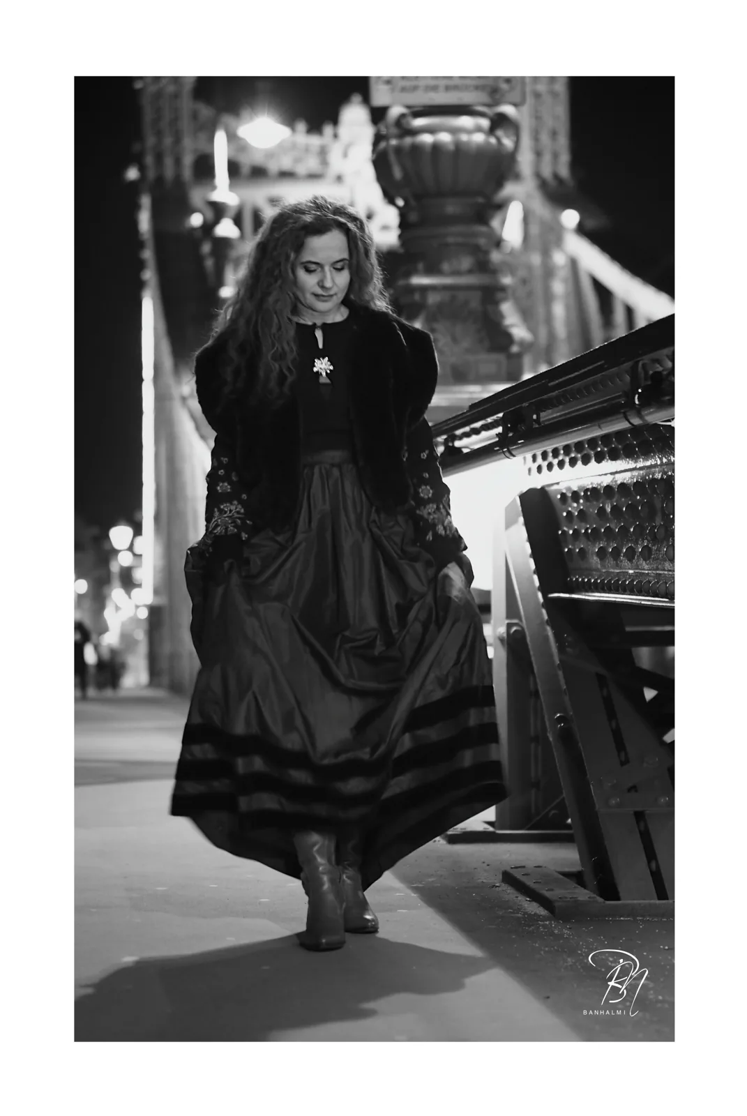 Night on the bridge — Photo: Norbert Banhalmi A woman in a long dark dress walks along a lit bridge at night.
