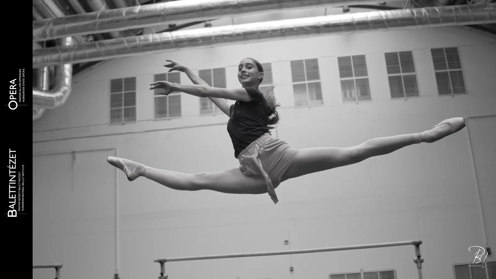 Grand jeté indoors — Photo: Norbert Banhalmi Black and white: a dancer in mid-air, legs fully extended in a grand jeté.