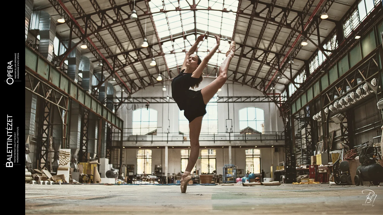 Under the iron roof — Photo: Norbert Banhalmi A dancer holds a high extension under the iron roof structure of a workshop hall.